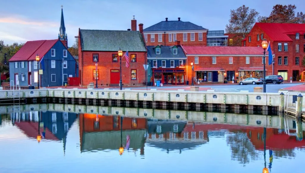 View of colorful blue and red Colonial-style homes near the water in Annapolis. The homes are reflected in the water in the foreground. 