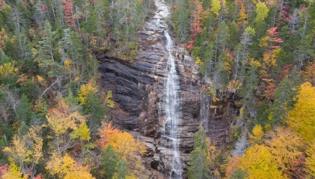 Aerial view of Arethusa Galls tumbling down a sheer cliff. It is surrounded by tall pine trees and trees turning yellow because of Autumn. 