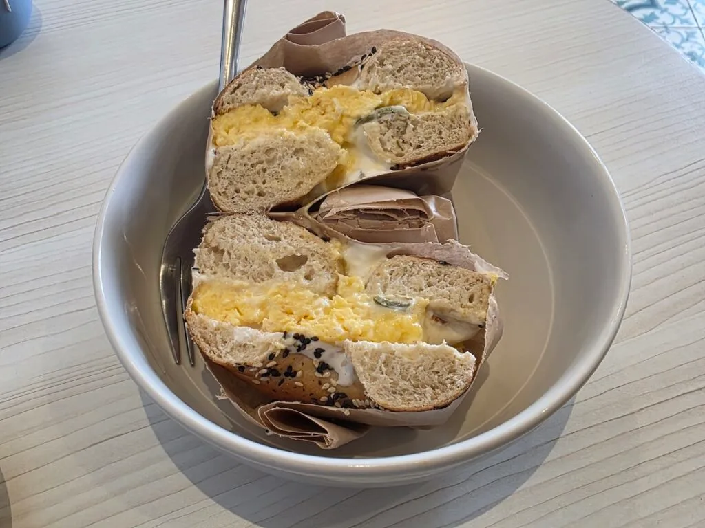 Aerial view of a bagel filled with eggs and jalapeno cream cheese. You can see a brown napkin around the bagel with a fork and in a white bowl. 