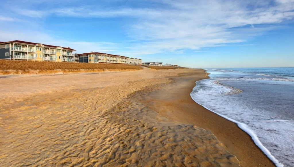 View of the golden beach with waves coming up against the shore with buildings in the background along the sand. 