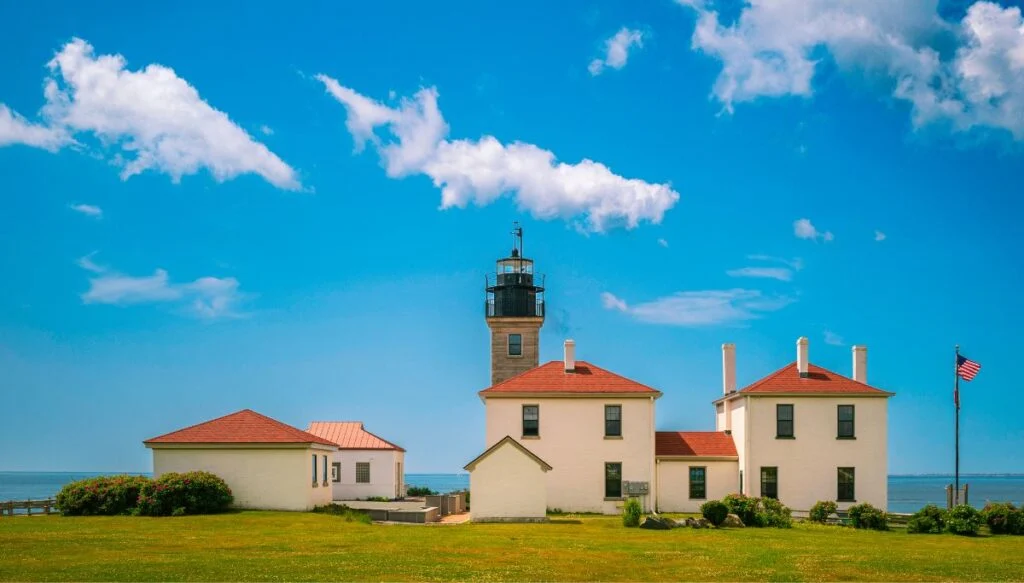 View of several white buildings sitting in front of Beavertail Lighthouse with the blue sky in the background and the American flag on the right on a sunny day. 
