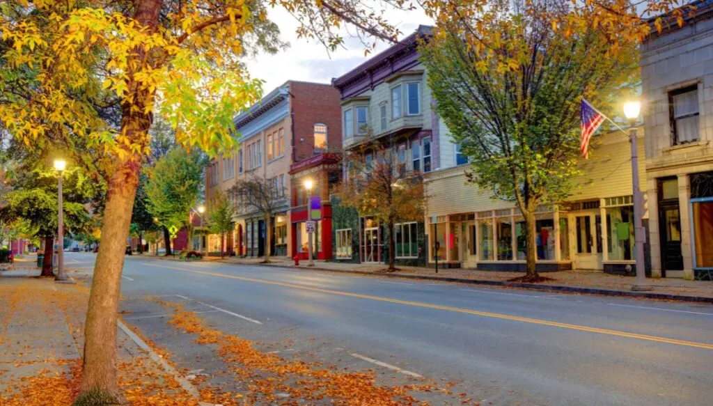 View of charming, old-world style stores lining the main street in Bennington at dusk. The lights are coming on and you can see vibrant foliage in the trees. 