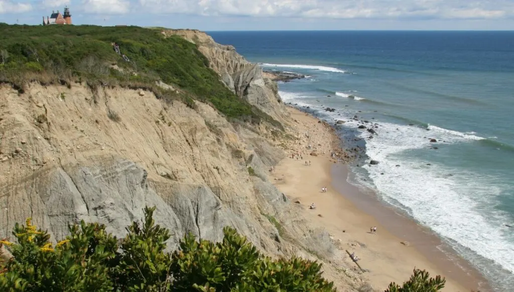View of the sheer rock faces of Mohegan Bluffs along the coast of Blok Island in Rhode Island. You can see the ocean along the edge of the cliffs and a lighthouse on the top of the cliff in the background. 
