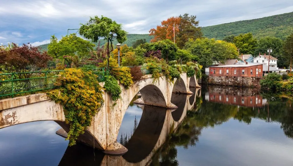 View of an arched white bridge covered in plants that extends across the river. You can see buildings in the distance and green mountains in the background. This is easily one of the most beautiful places in Massachusetts. 