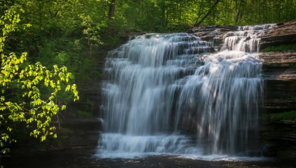 View of Buttermilk Falls tumbling down a series of rocks and surrounded on the left by a ton of green trees. This is easily one of the most beautiful places in New York state. 