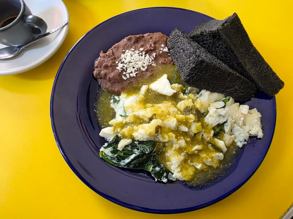 Aerial view of eggs with a yellow sauce and spinach with beans next to it and black bread on the side with an espresso on a yellow table at Cafe Regina. 