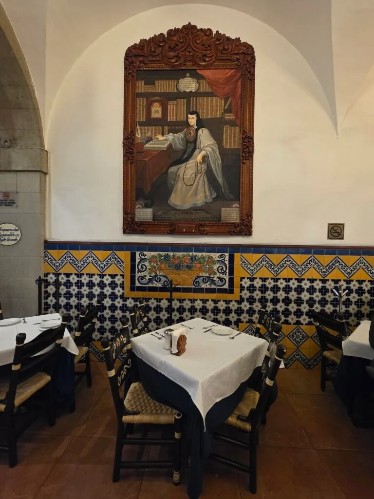 View of a painting of a man inside of Cafe de Tacuba, You can see bright litles beneath the painting and tables in the dining rooms