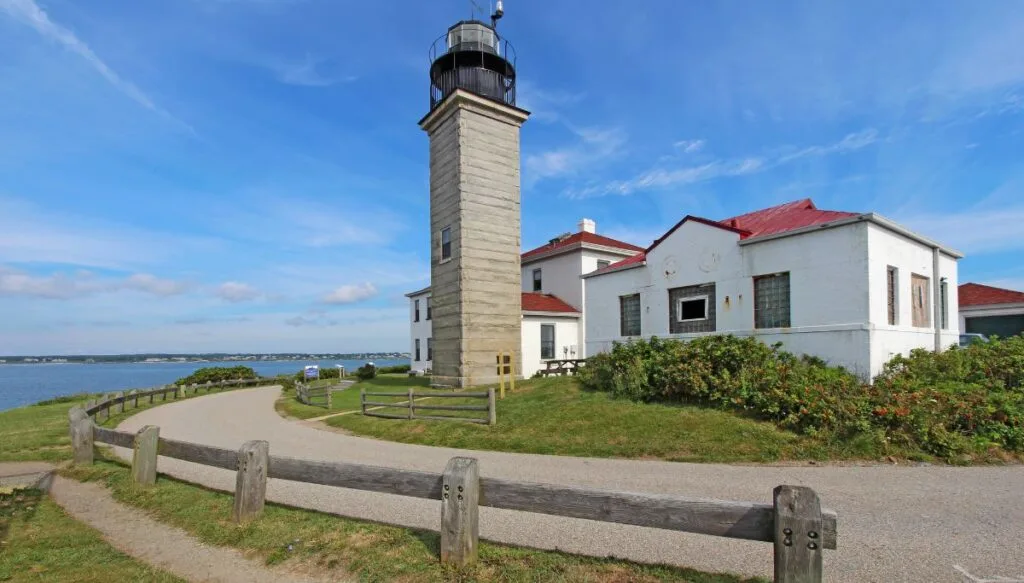 View of a small lighthouse with a gray stone tower and a white house behind it. In the front you can see a paved walking path that takes you along the water. 