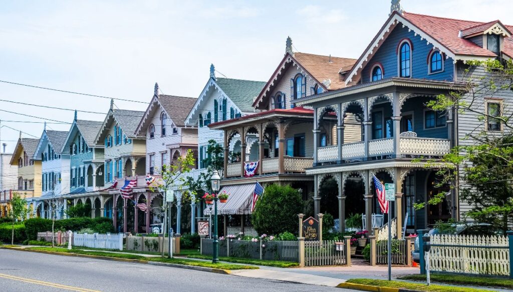 View of a row of homes sitting along a street in Cape May. The homes are Victorian-style and come in a vriety of colors with a double decker porch in the front and American flags.