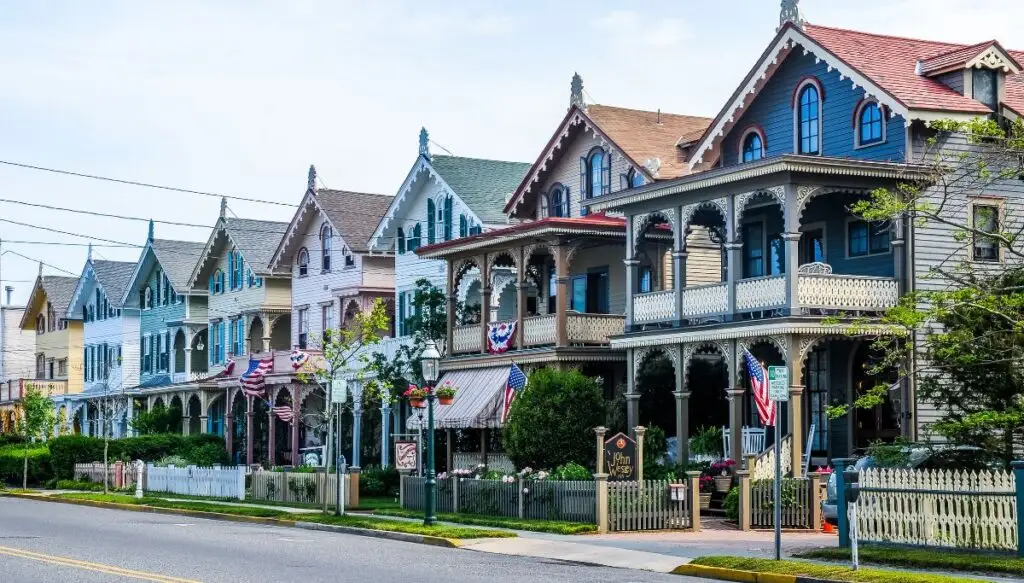 View of a row of homes sitting along a street in Cap May. The homes are Victorian-style and come in a vriety of colors with a double decker porch in the front and American flags. 