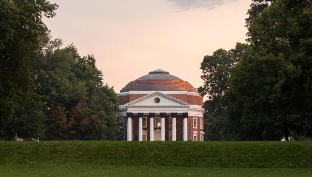 View of the Rotunda at the University of Virginia. They sky is darkening and the building is surrounded by trees with a domed roof and columns out front. 