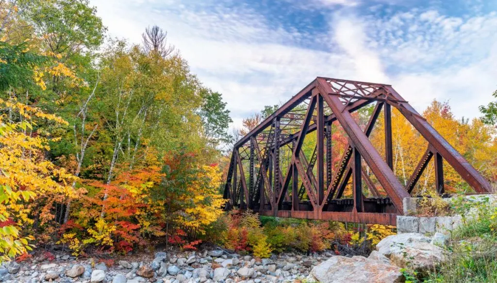 View of a rusted stainless steel bridge over a dry river with bright orange and yellow and green trees in Crawford Notch State Park. 