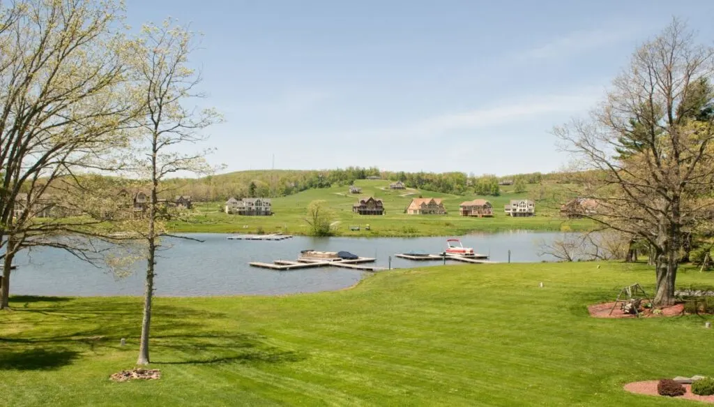 View of grassy fields surrounding a lake with houses around it and piers in the water. You can also see trees in the foreground. 