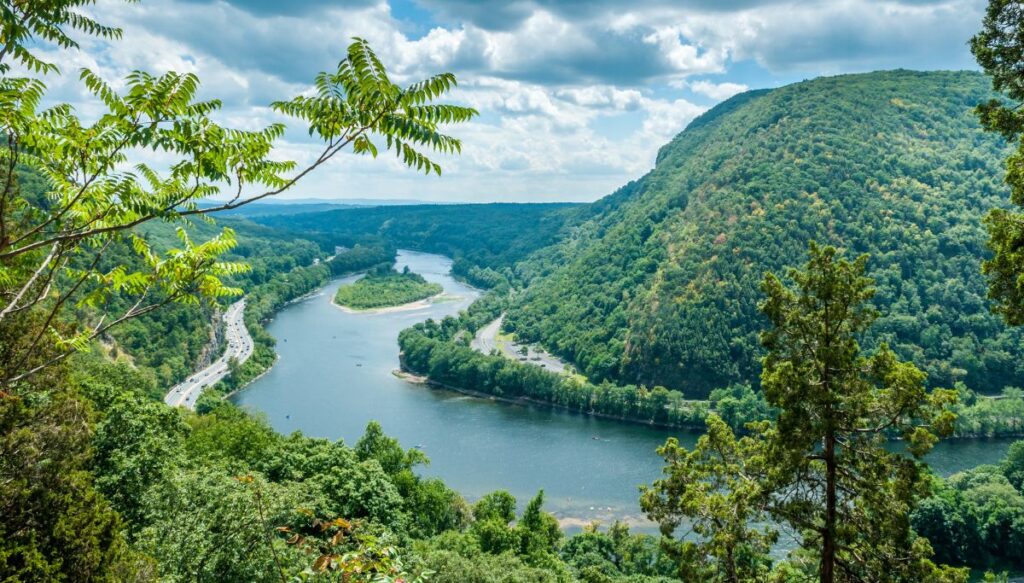View of the lush green mountains surrounded a river at the Delaware Water Gap on a cloudy day with some trees in the foreground.