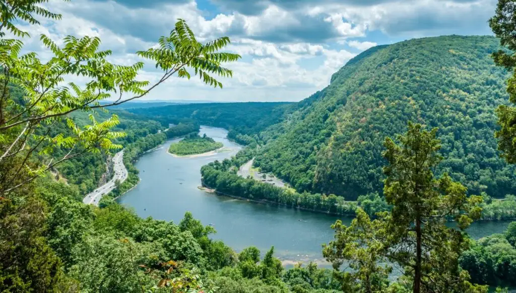 View of the lush green mountains surrounded a river at the Delaware Water Gap on a cloudy day with some trees in the foreground. 