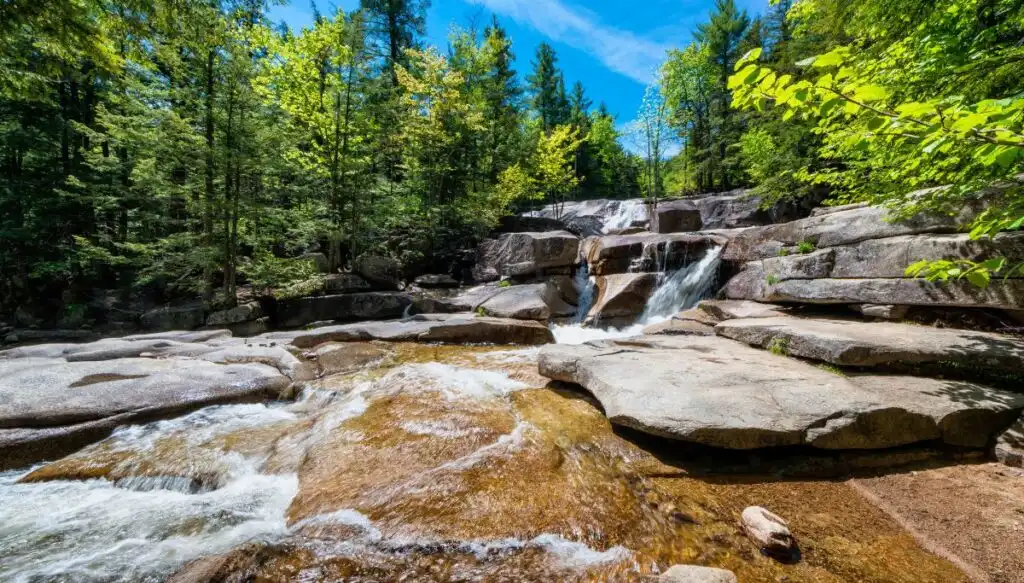 View of a series of small waterfalls tumbling down large gray rocks with green trees all around it at Diana's Baths in New Hampshire. 