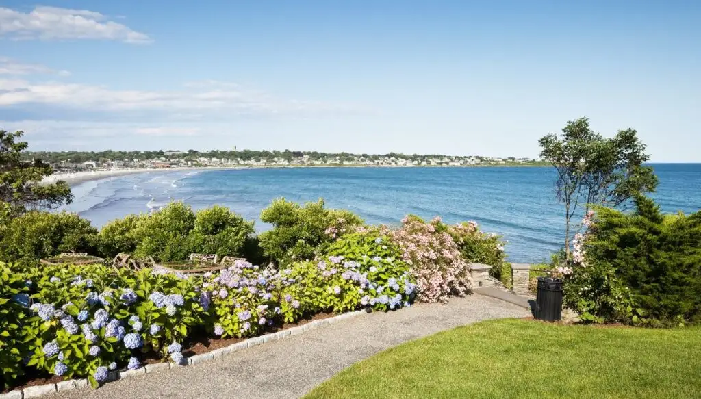 View of a park with a paved path with beautiful flowers on the left. You can see the water in the distance and Easton'a Beach in the distance on the left. 