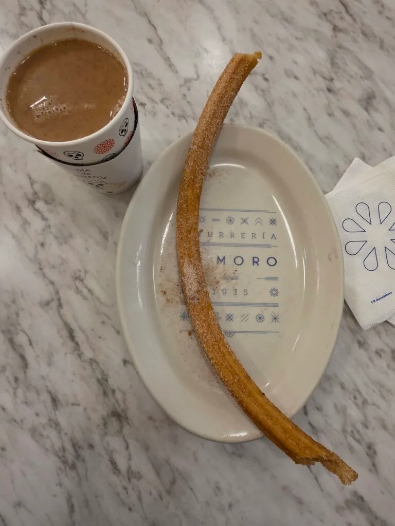 Aerial view of a Churro sitting on a white plate with a hot chocolate on the left and it is sitting on a marble countertop. 