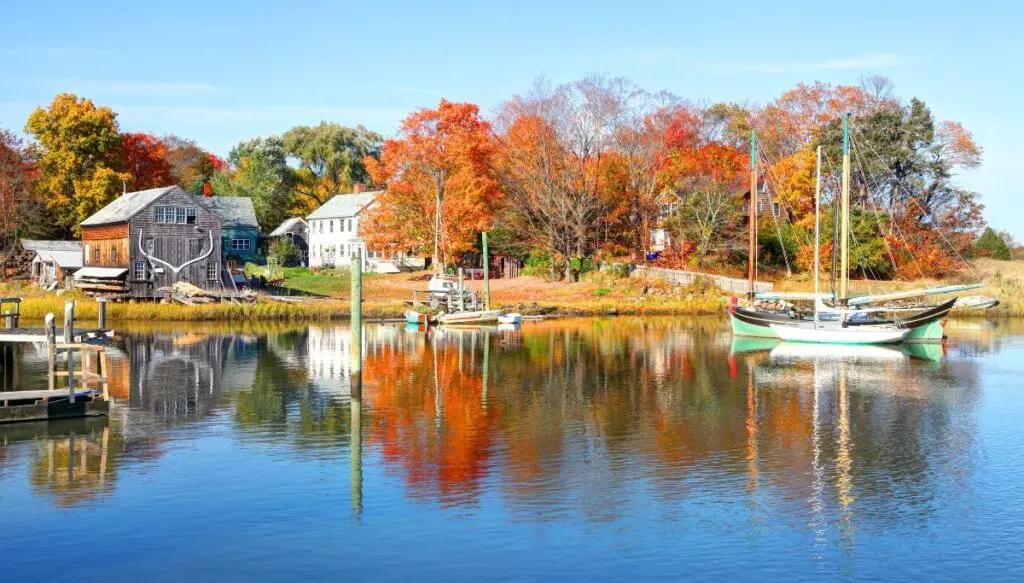 View of houses sitting amidst fall foliage along the water with sailboats bobbing in the water. 