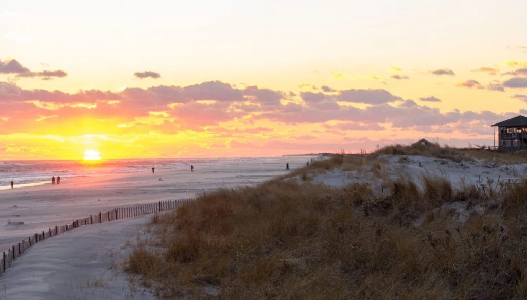 View of the sun rising above the beach at Fire island seashore. You can see people walking along the beach and grass covered dunes in the foregrounf with a building on the right. 