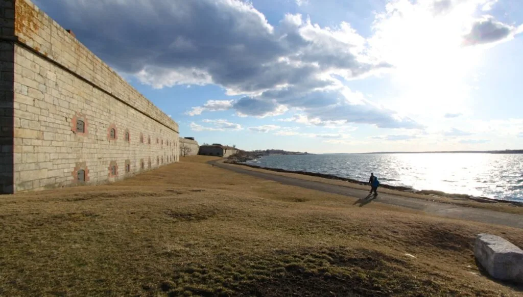 View of the stone wall of a fort on the left and a walkway with people on it on the right that takes them along the ocean. 