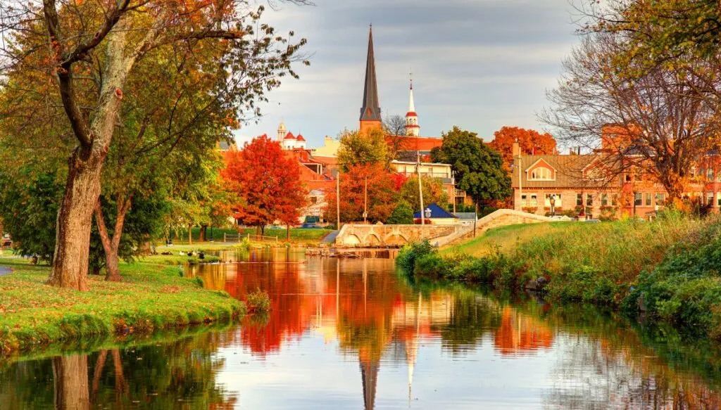 View of a creek surrounded by trees with vibrant fall foliage in the background and historic buildings in the distance in Frederick. This is one of the most beautiful places in Maryland. 