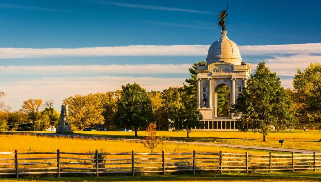 View of the Pennsylvania Monument in Gettysburg. The sun is setting and the grass looks yellow with tall trees and a wooden fence in the foreground. You can also see a stone monument in the background. 