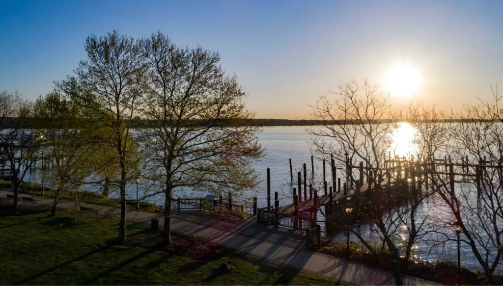 View of trees with some leaves on the grass with a paved path in front near the water with a wooden dock going out into the water in Havre d Grace. You can also see the sun getting lower in the sky. 