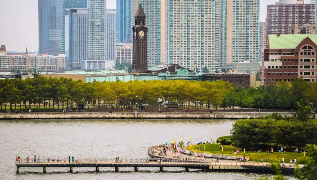 View of the skyline of Jersey City with a walking path along the water and a clock tower in the distance. 