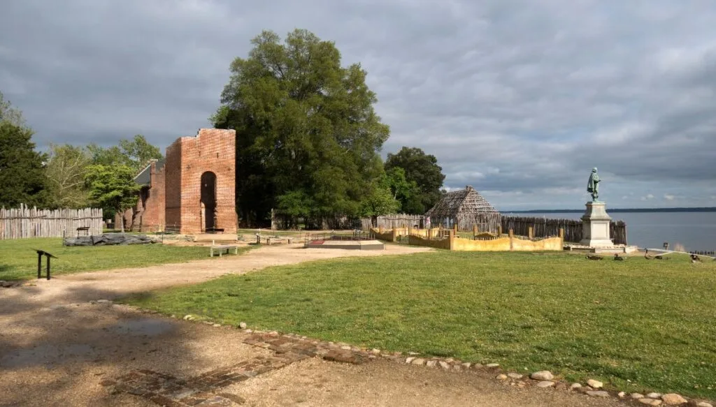 View of the ruins inside of Jamestown. You can dirt pathways leading to an old brick building and w wooden structure that sits next to the river. 