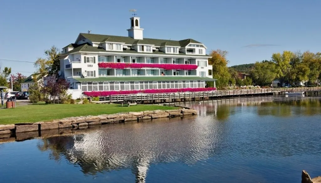 View of a large white hotel with balconies overlooking the edge of  Lake Winnipesaukee on a sunny day. 