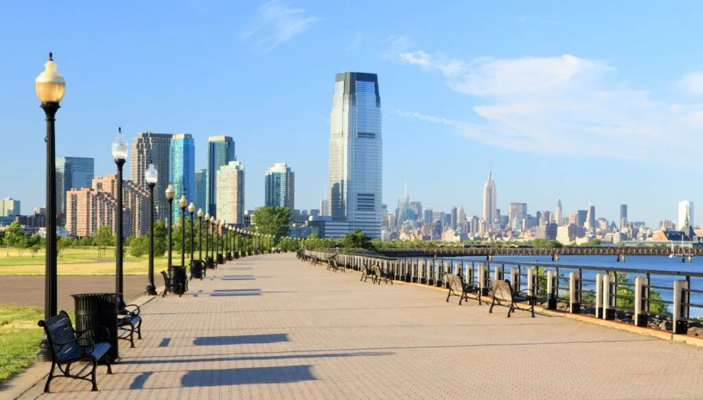 View of the boardwalk along the river in Liberty State Park. Benches and light posts line the riverside walkway and you can see skyscrapers in the background. 