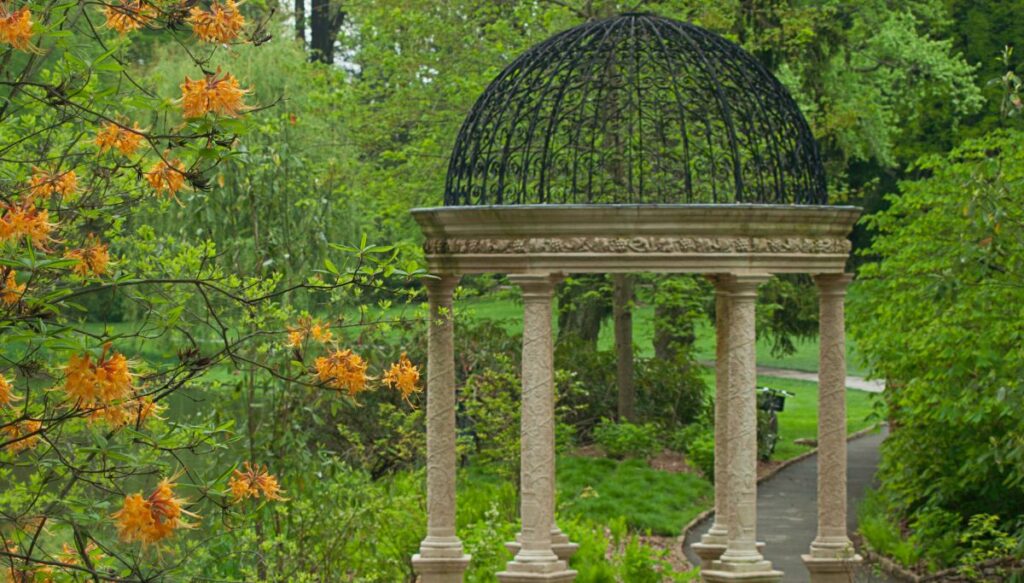View of a pergola with marble columns and a metal roof. You can see bright orange flowers to the left and a walking path going past trees inside Longwood Gardens. This is one of the most beautiful places in Pennsylvania.
