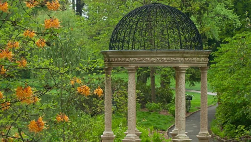 View of a pergola with marble columns and a metal roof. You can see bright orange flowers to the left and a walking path going past trees inside Longwood Gardens. This is one of the most beautiful places in Pennsylvania. 