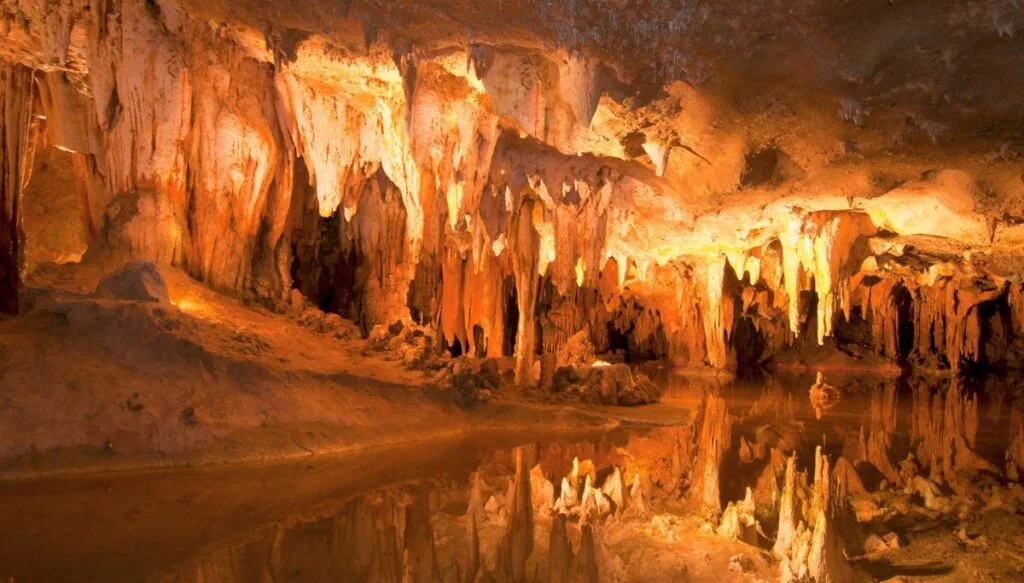 View of a white light illuminating the rock formations inside of Luray Caverns with water on the ground. 