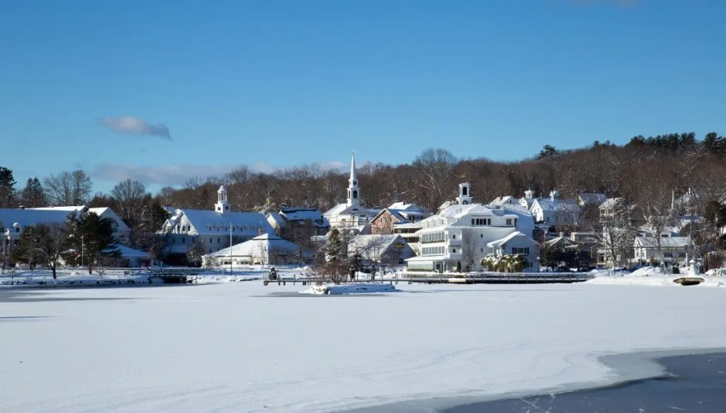 View of an assortment of buildings covered in snow in the winter. You can see a frozen lake in the foreground and buildings in the background. 