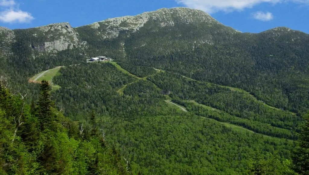 View of Mount Mansfield in the distance covered in pine forests with a small lodge near the top in an open area. 