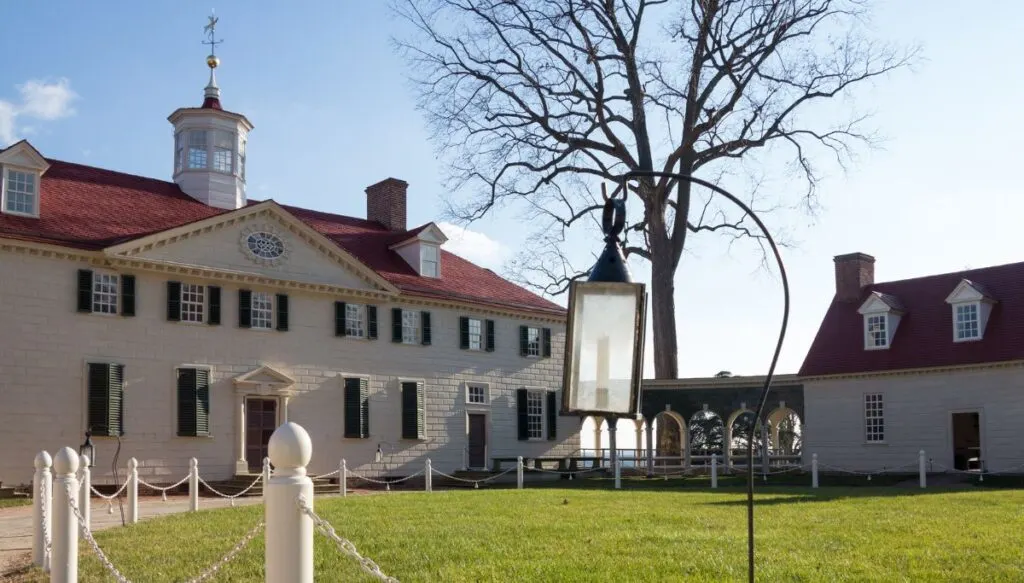 View of the white Colonial-style buildings that make up Mount Vernon. You can see a red rood and black shutters and a road in front of the building. 