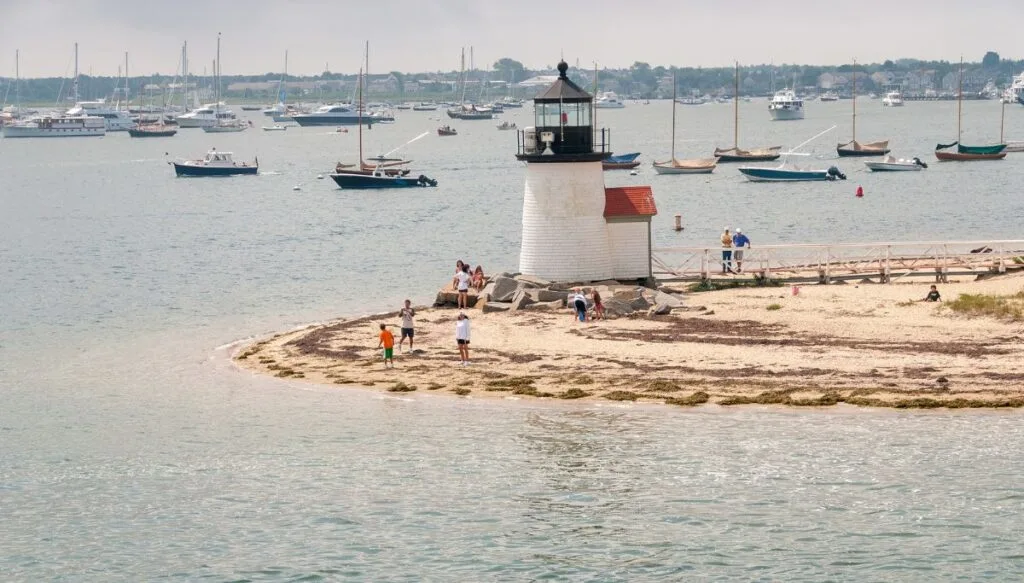 Aerial view of a small white lighthouse with people walking on the beach around it. You can see sailboats in the distance in Nantucket. 