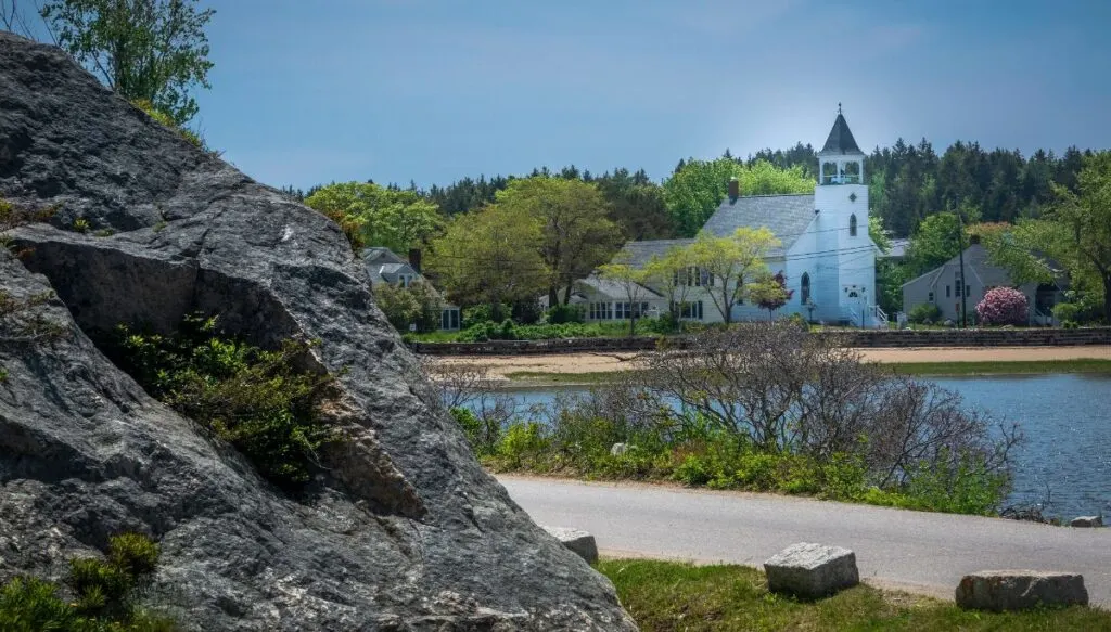 View of a rock in the foreground on the left and a road behind it with a beach in the distance with a white church behind it at Popham Beach State Park. 