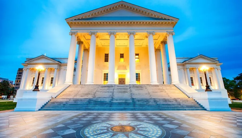 View of the white state capitol with columns out front all lit up in the evening in Richmond. 