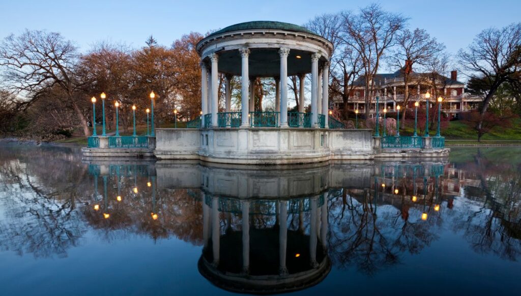 View of a stone, circuliar pavillion sitting along the water with lights around it and large mansions in the background. This is inside Roger Williams Park and its one of the most beautiful places in Rhode Island.