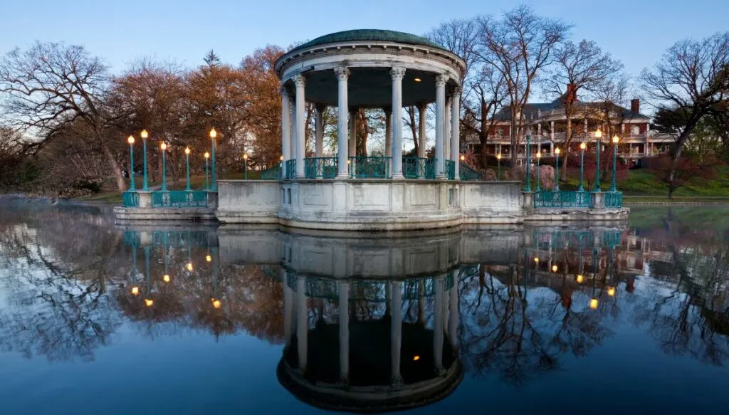View of a stone, circuliar pavillion sitting along the water with lights around it and large mansions in the background. This is inside Roger Williams Park and its one of the most beautiful places in Rhode Island. 