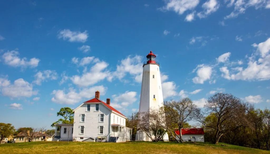 View of a white lighthouse with a red top and a white houses with a red roof next to it in Sandy Hook. 
