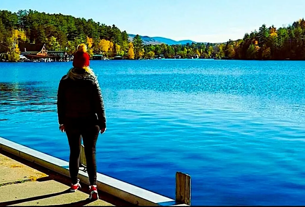 View of me standing on a pier on the edge of saranac Lake in a black jacket and red hat with foliage along the edge of the lake. 