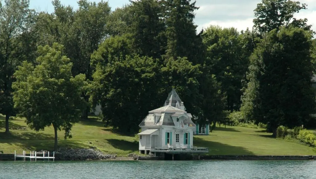 View of a white, Colonial-style boathouse sitting along the edge of Skaneateles Lake with giant pine trees all around it. 