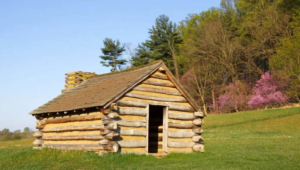 View of a charming log cabin sitting in a field with trees behind it and blooming pink flowers at Valley Forge. 