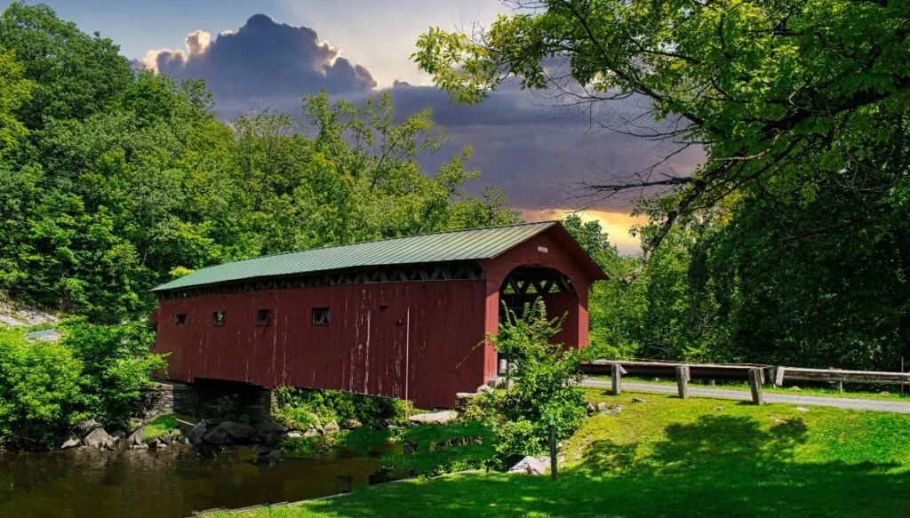 View of a red covered bridge going across a river surrounded by greenery with clouds in the background. 