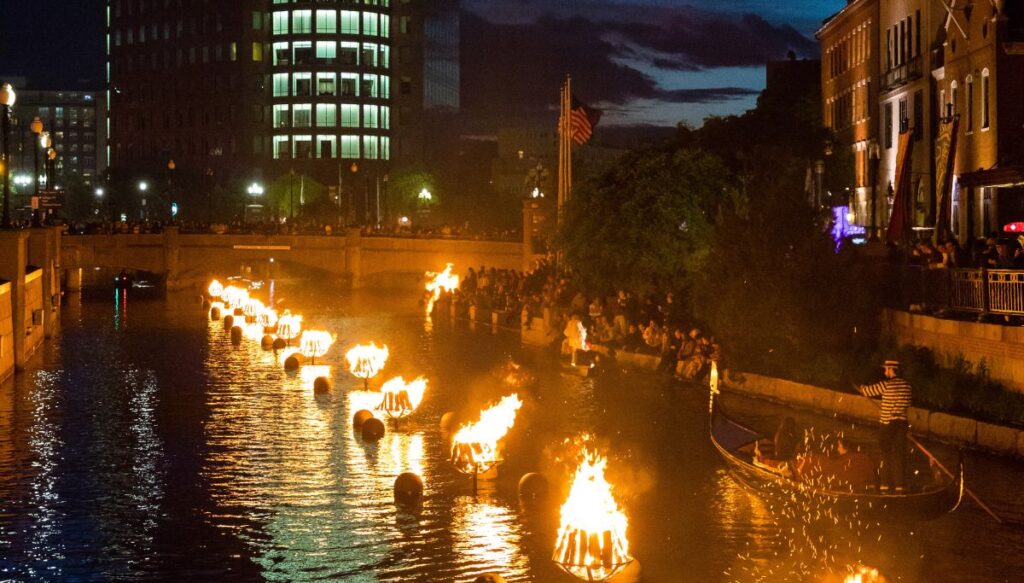 View of bon fires all lit up along the river in Providence with people standing in the background and skyscrapers in the back at night.