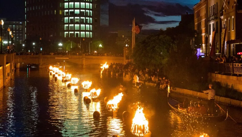 View of bon fires all lit up along the river in Providence with people standing in the background and skyscrapers in the back at night. 